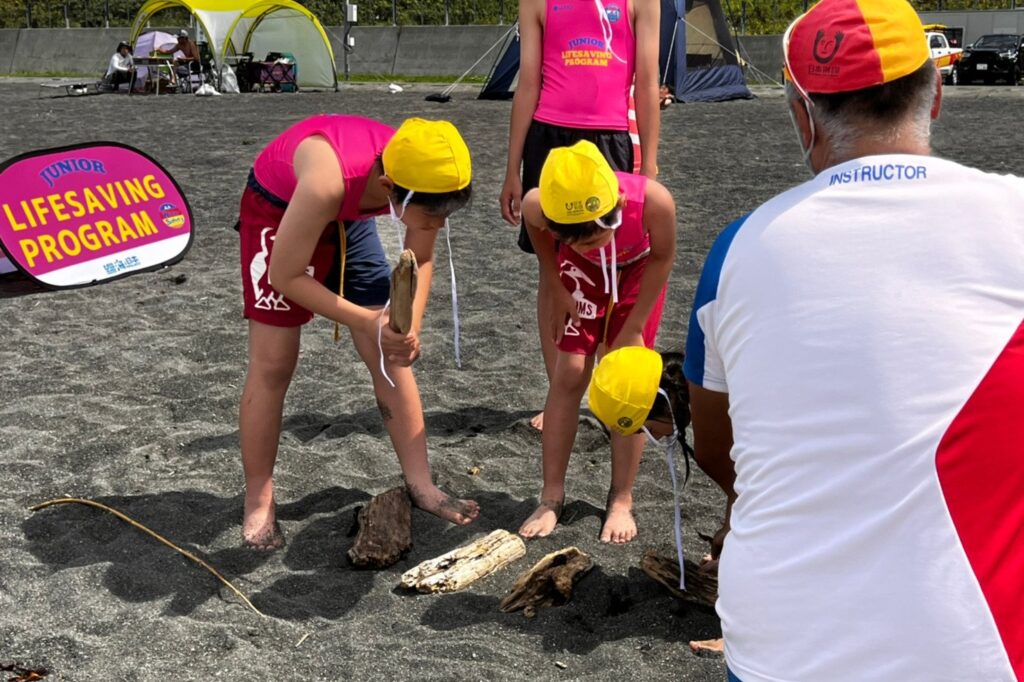 砂浜で見つけた流木を並べ、かがんでのぞきこむ子どもたちとインストラクター