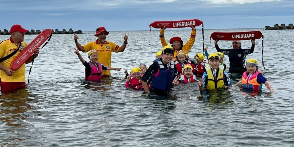 腰までの海に入り、レスキューチューブを頭の上に掲げるライフセーバーと、ライフジャケット姿で手を上げる子どもたち