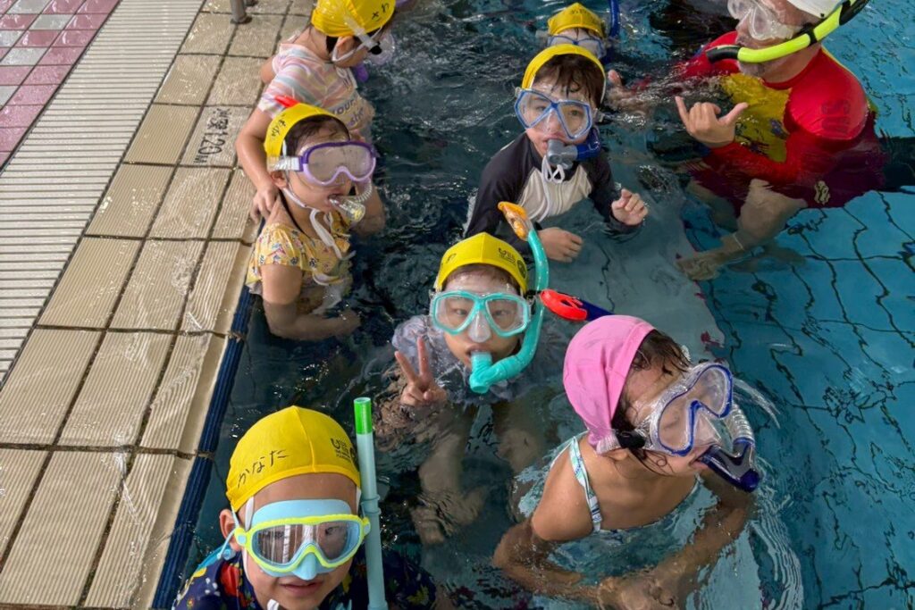 プールでマスクとシュノーケルをつけて、カメラに向かってピースする子どもたち