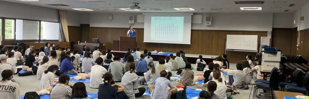 大人数の会場で行った救命講習。訓練用人形を前に、前方のスクリーンで説明を聞く参加者