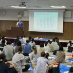 大人数の会場で行った救命講習。訓練用人形を前に、前方のスクリーンで説明を聞く参加者